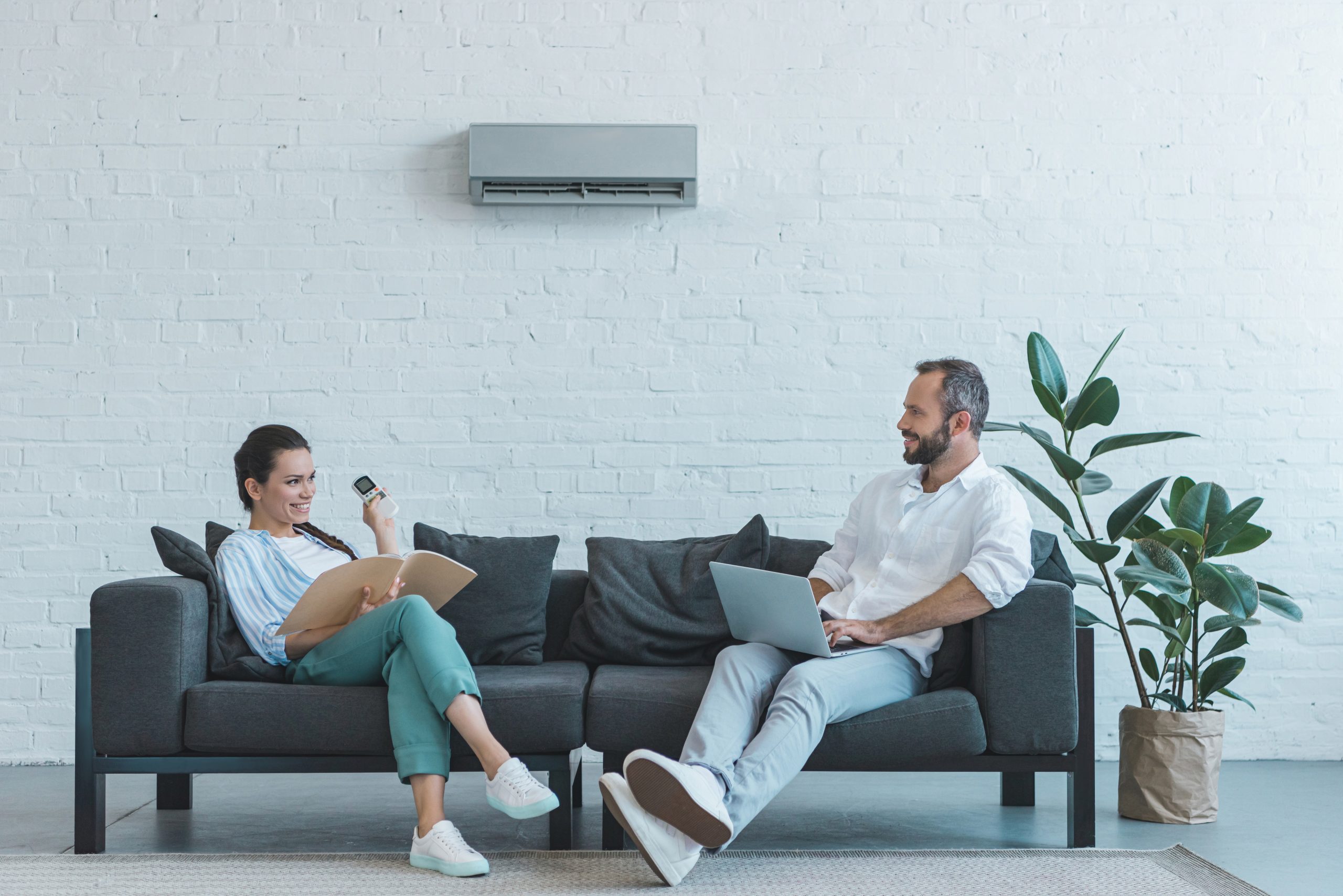 Couple turning on air conditioner during the summer heat while sitting on sofa with book and laptop